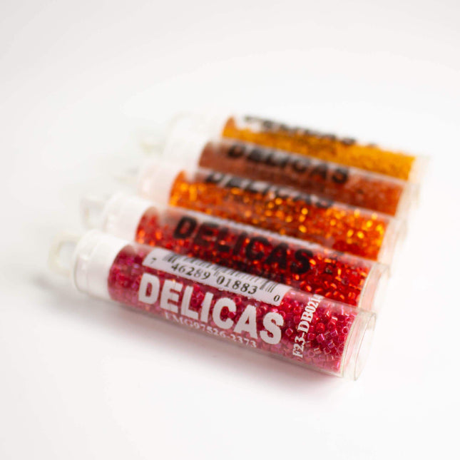 a collection of colorful glass beads, each with a label that reads "DELICAS". The beads are arranged in a row, with the first one being red, the second one orange, the third one yellow, and the fourth one pink.