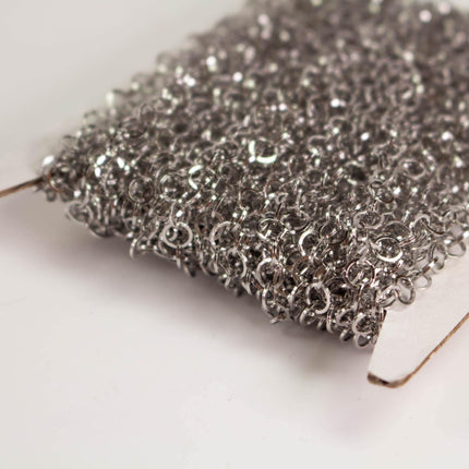 A close-up image of a pile of silver-colored metal rings or links, creating a textured and shiny appearance.